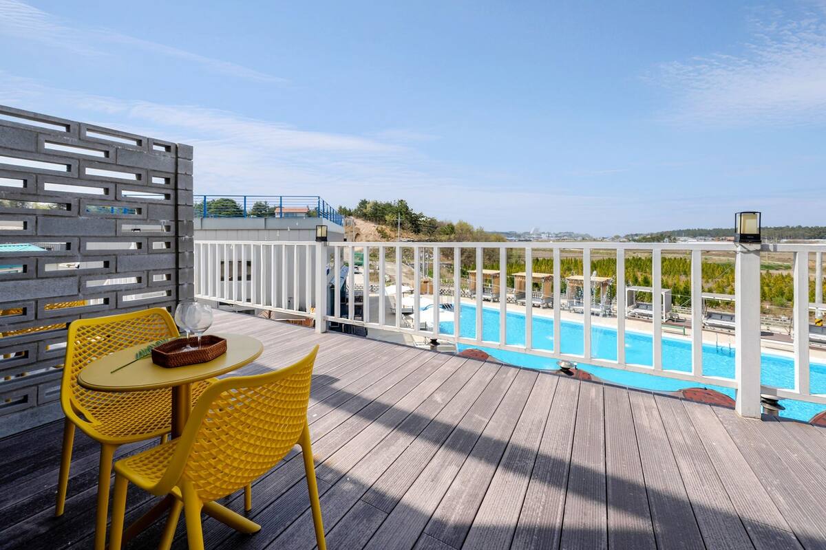 A wooden deck with yellow chairs and a table, overlooking a swimming pool with a view of hills and a distant building.