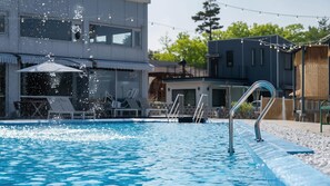 Een zwembad met helder water, omringd door moderne gebouwen, een loungegedeelte met parasols, en een lichtslingering.