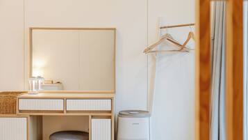 A hotel room with a wooden vanity, a mirror, a hanging clothes rack, a trash bin, and a window with curtains.