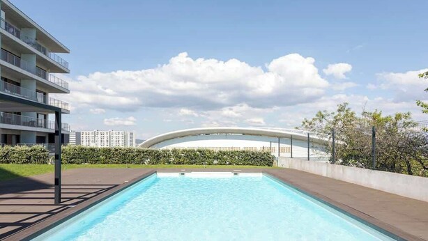 Una piscina rectangular con superficie de agua azul cristalina, rodeada por un muro de ladrillo bajo y una barandilla metálica. Al fondo, se observa un edificio moderno con techo curvo y fachada blanca.