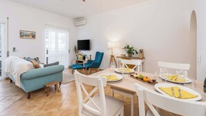 Una habitación luminosa con una mesa de comedor para dos personas, un sofá, un televisor y un ventilador de techo.