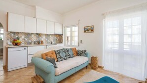 Un espacio habitable compacto con kitchenette, un sofá, un taburete pequeño y una ventana con cortinas traslúcidas.