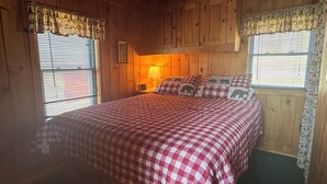 A wooden cabin room with a bed, two windows with curtains, and a lamp. - Idlewood Cabin 3 (Rangeley)