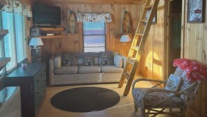 A cozy cabin interior with a wooden ceiling, a sofa, a chair, a TV, and a ladder. - Idlewood Cabin 3 (Rangeley)
