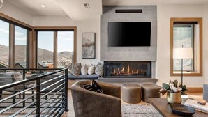 A modern living room with a fireplace, a large flat-screen TV mounted on a wall, a balcony with a railing, and a view of the landscape.