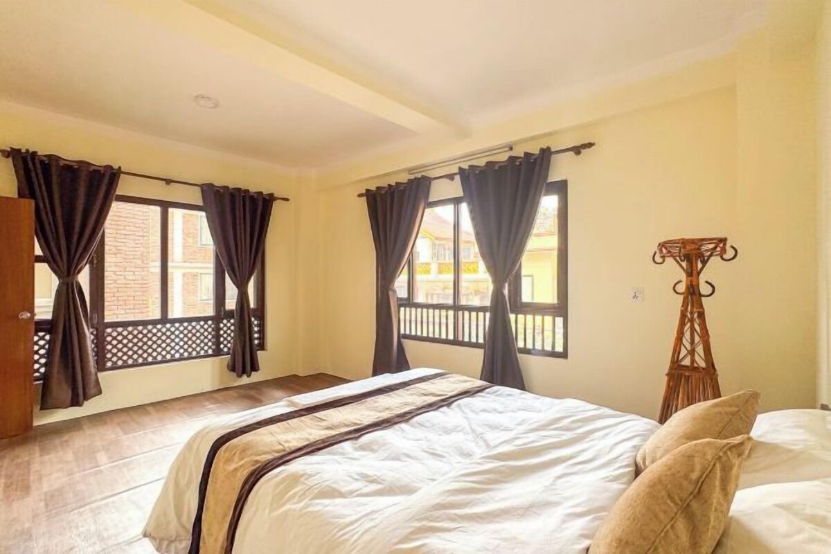 A bedroom with a bed, curtains, a wooden floor, and a window with a view of buildings outside.