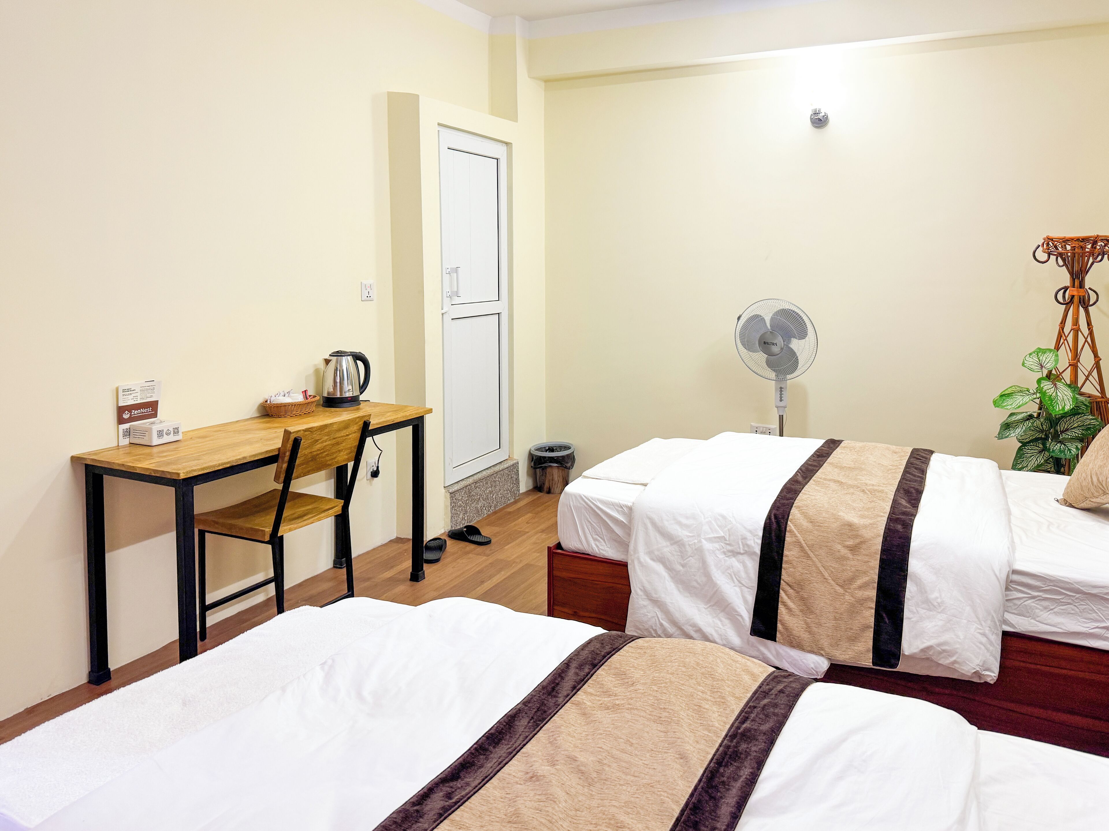 A hotel room with two beds, a desk, a chair, a fan, a plant, and a door.