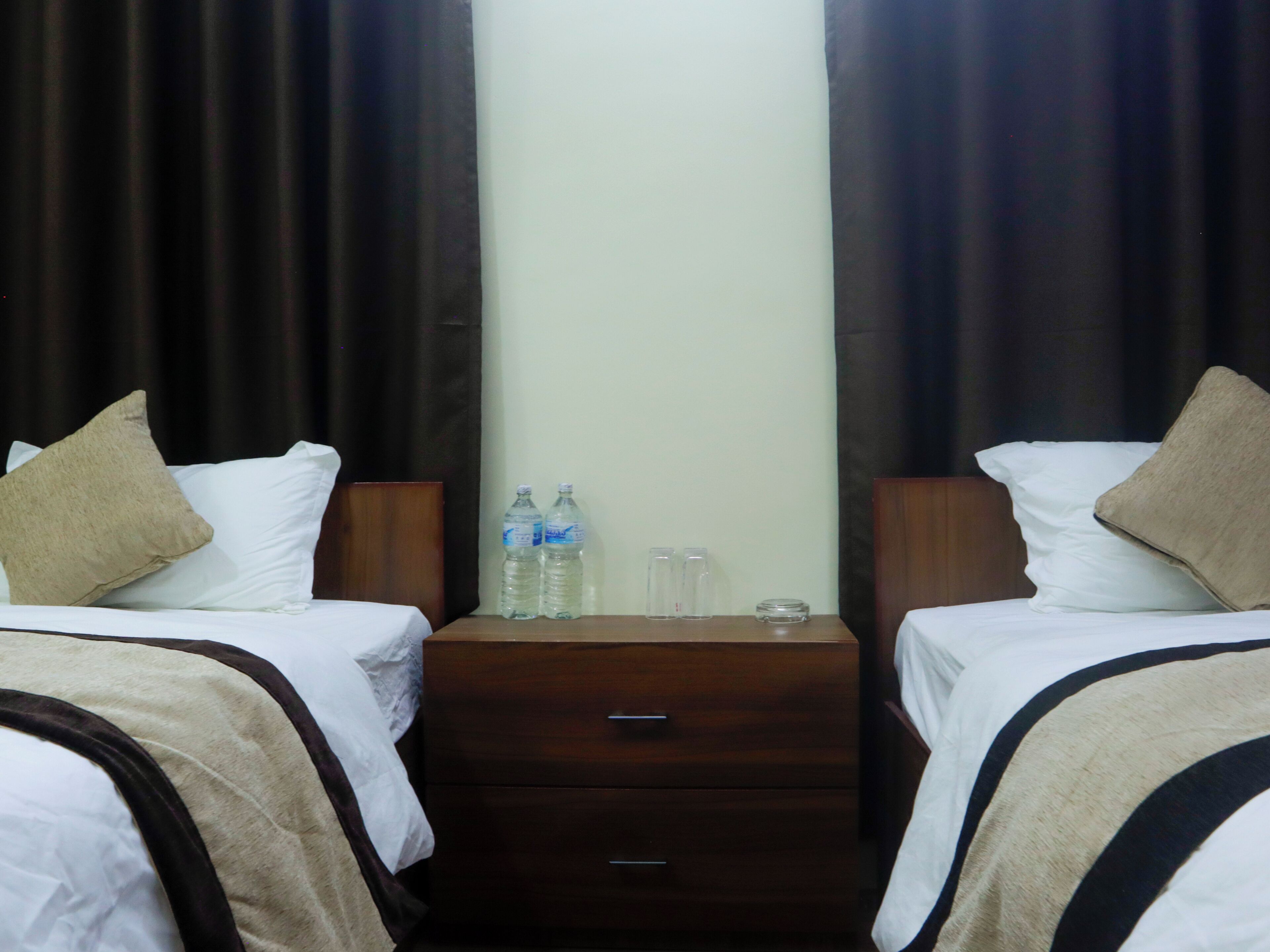 A hotel room with two beds, each with a pillow, a wooden nightstand with two bottles of water and two glasses, and a small drawer.