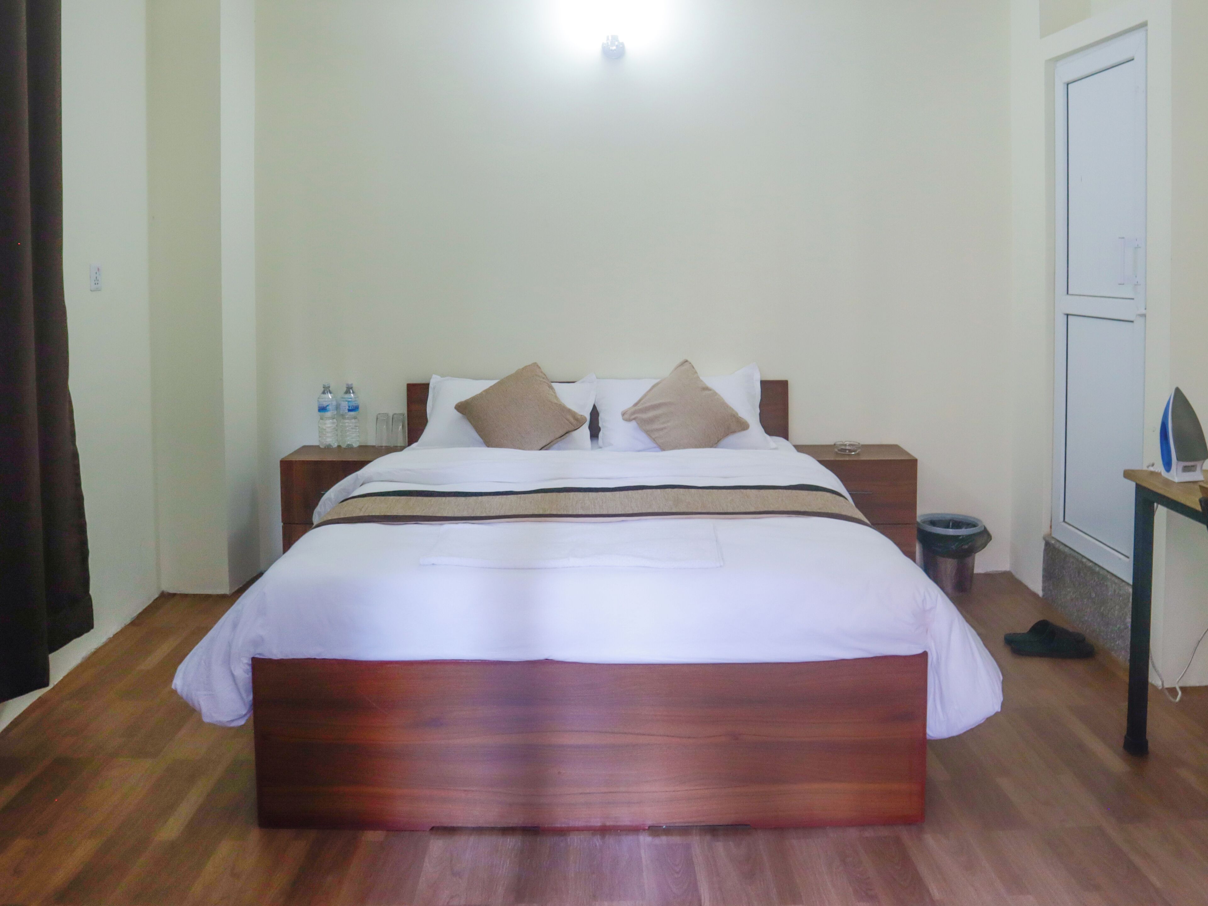 Una cama bien hecha con cabecera de madera, una mesita de noche con lámpara y un cubo de basura.