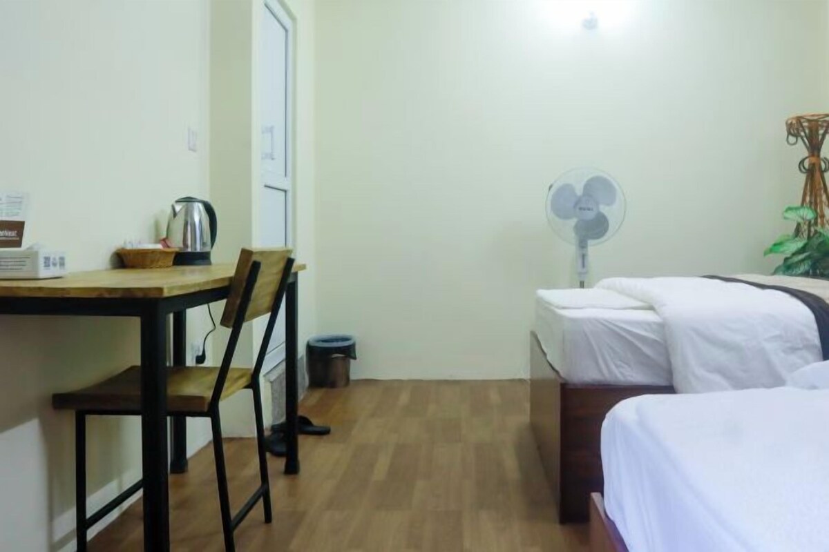 A small, clean room with a wooden desk, a chair, a bed with white bedding, a standing fan, and a small trash bin.
