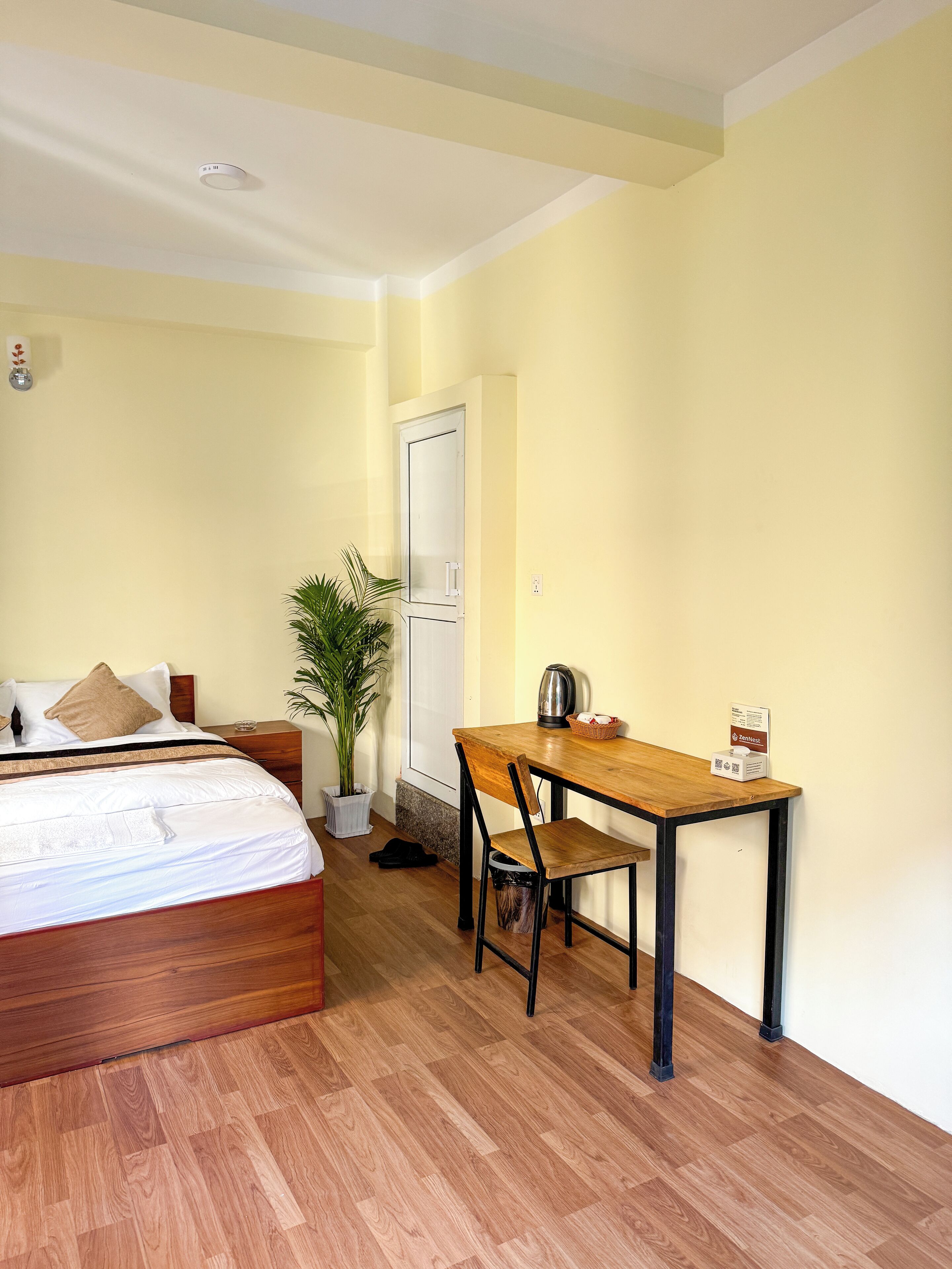 A hotel room with a bed, a wooden desk, a chair, a potted plant, and a door.
