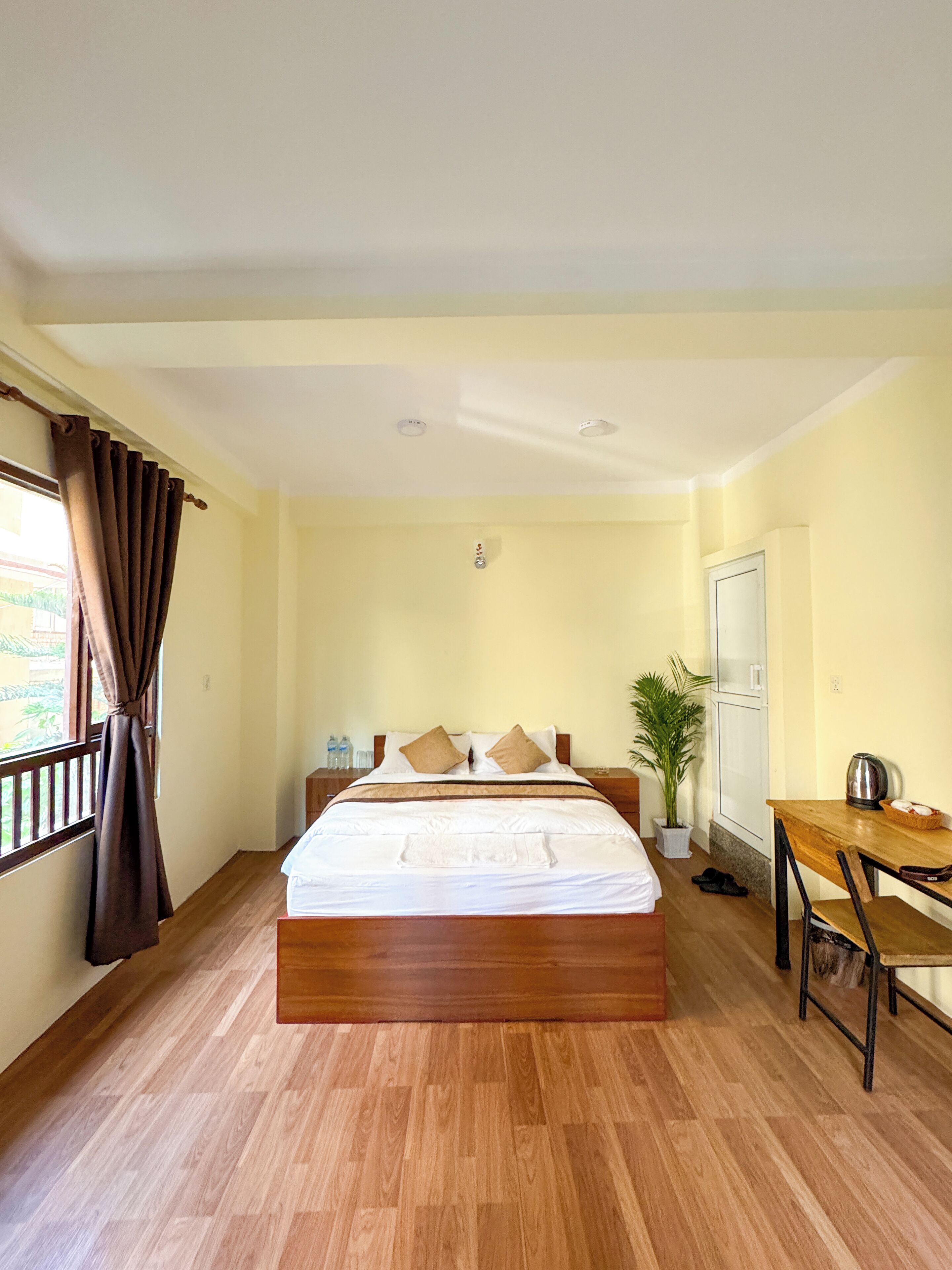 A hotel room with a wooden bed, a desk, a potted plant, and a view of the outdoors.