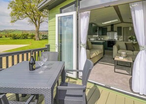 Un balcon avec une table et des chaises, une porte-fenêtre coulissante donnant sur un espace de vie comprenant un canapé et une petite table, et une vue sur un paysage verdoyant.
