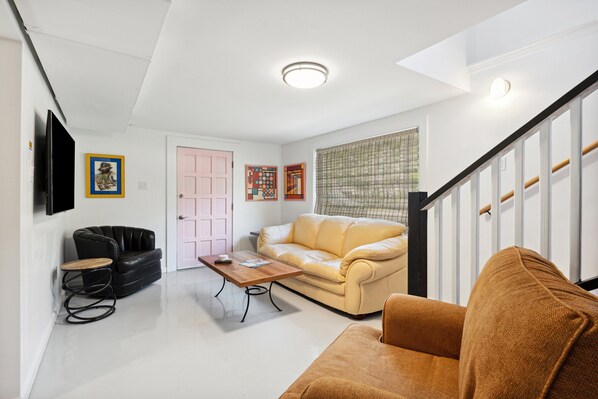 A living room with a beige sofa, a black leather chair, a wooden coffee table, and a brown armchair. There is a pink door and a framed artwork on the wall.