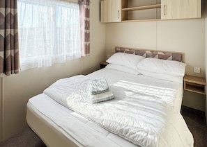 Un lit bien fait, avec du linge de lit blanc, une tête de lit ornée d’un motif géométrique, une table de chevet et une fenêtre avec des rideaux légers.