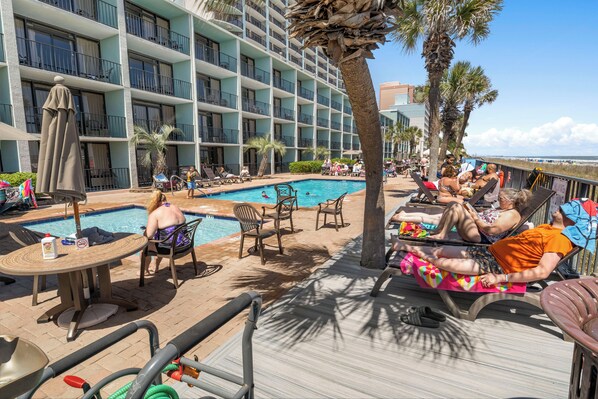A poolside area with lounge chairs, a table, and a view of a hotel building. - Oceanview Vibes! Compass Cove Studio Retreat (Myrtle Beach)