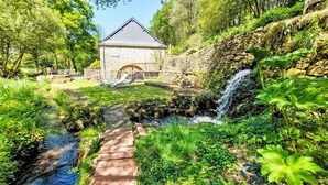 A stone building with a sloped roof, a small waterfall, and a wooden pathway leading to it.