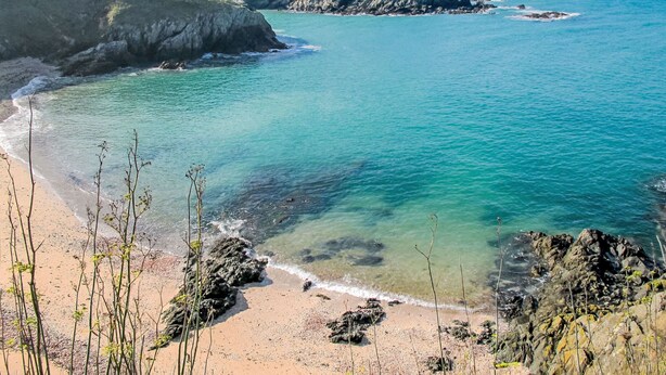 A coastal landscape with clear turquoise waters, rocky shores, and grassy hills.