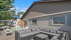 A covered outdoor seating area with a large sectional sofa and a small table, adjacent to a house with a sloped roof and large windows. - Little Sand Cottage 3-bedroom cottage with AC, WiFi in serene Grant (Grant)