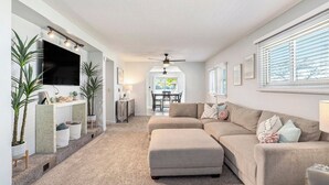 A spacious living room with a sectional sofa, a flat-screen TV mounted on the wall, and a ceiling fan. There are potted plants and a small table with a lamp. - Little Sand Cottage 3-bedroom cottage with AC, WiFi in serene Grant (Grant)