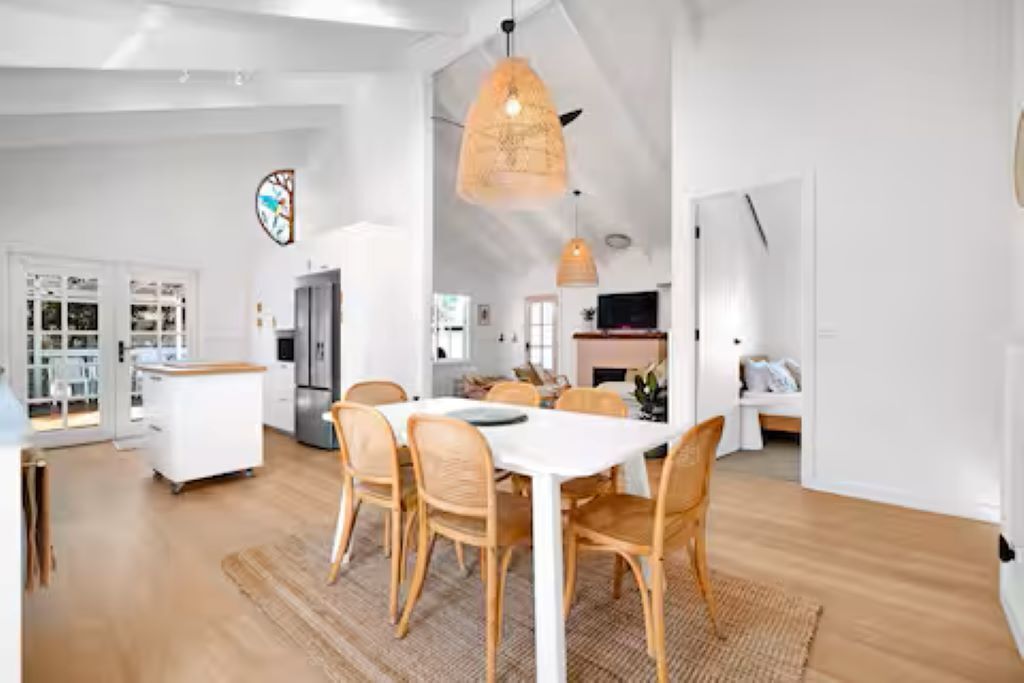 Un vaste espace de vie avec une grande table et des chaises blanches, un luminaire suspendu et, au fond, une vue sur la cuisine.