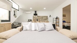 Une chambre moderne avec un grand lit, une commode en bois, un banc intégré et un coin cuisine à l’arrière-plan.