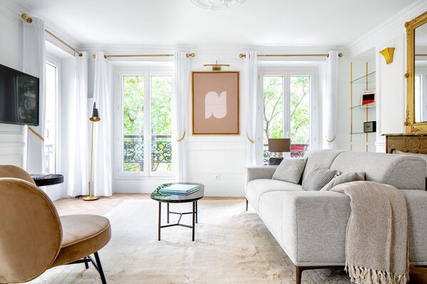 Una sala de estar con sofá, sillón y una mesita. Hay ventanas amplias con cortinas, un televisor y un cuadro en la pared.