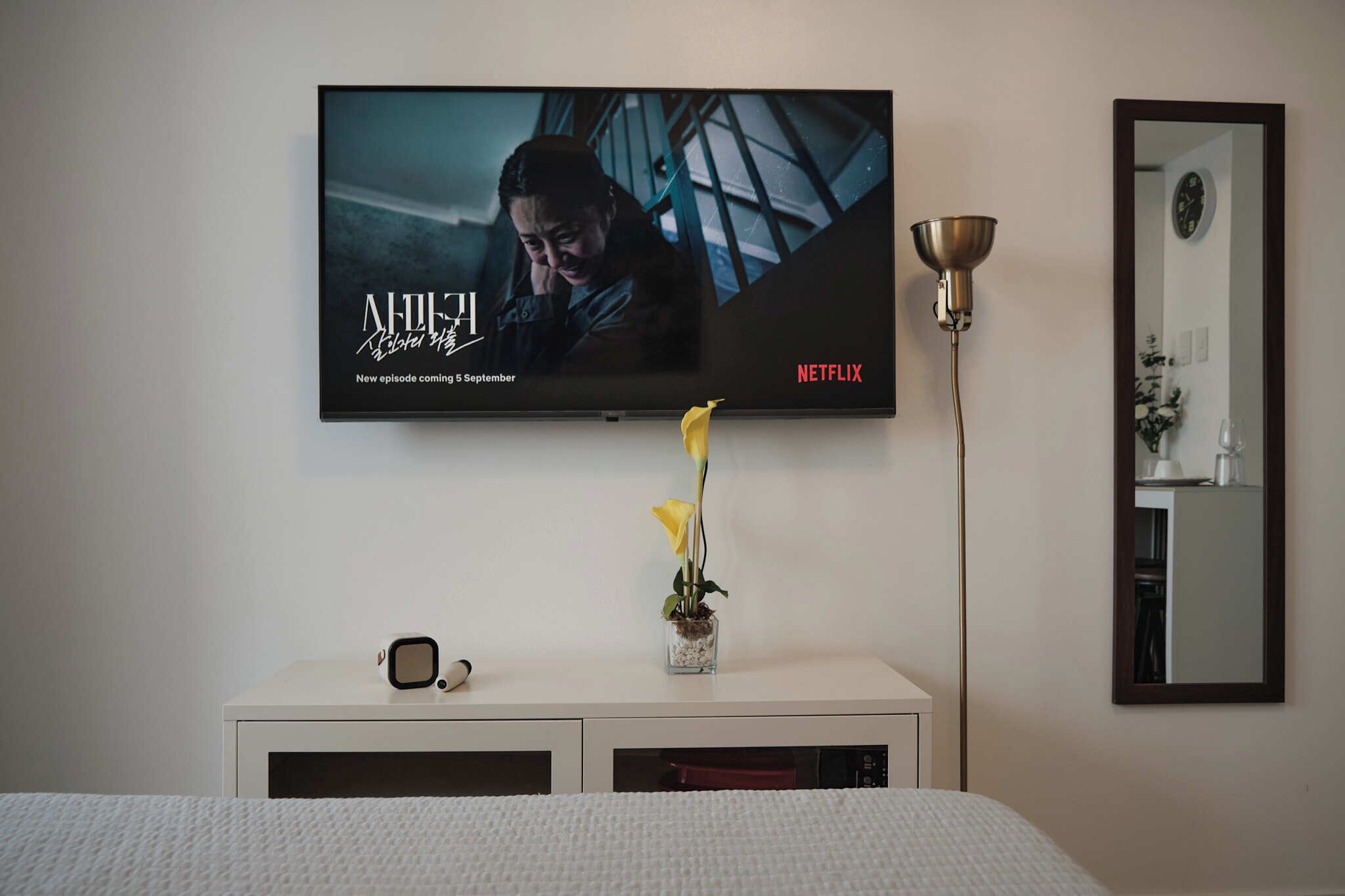 Un televisor montado en la pared mostrando una serie de Netflix, una mesita de noche con un florero y una lámpara de noche.