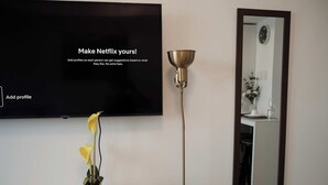 Un téléviseur à écran plat est fixé au mur et affiche l’interface de Netflix, avec un bouton intitulé « Ajouter un profil ». Une fleur jaune en pot est posée sur une table à côté du téléviseur.