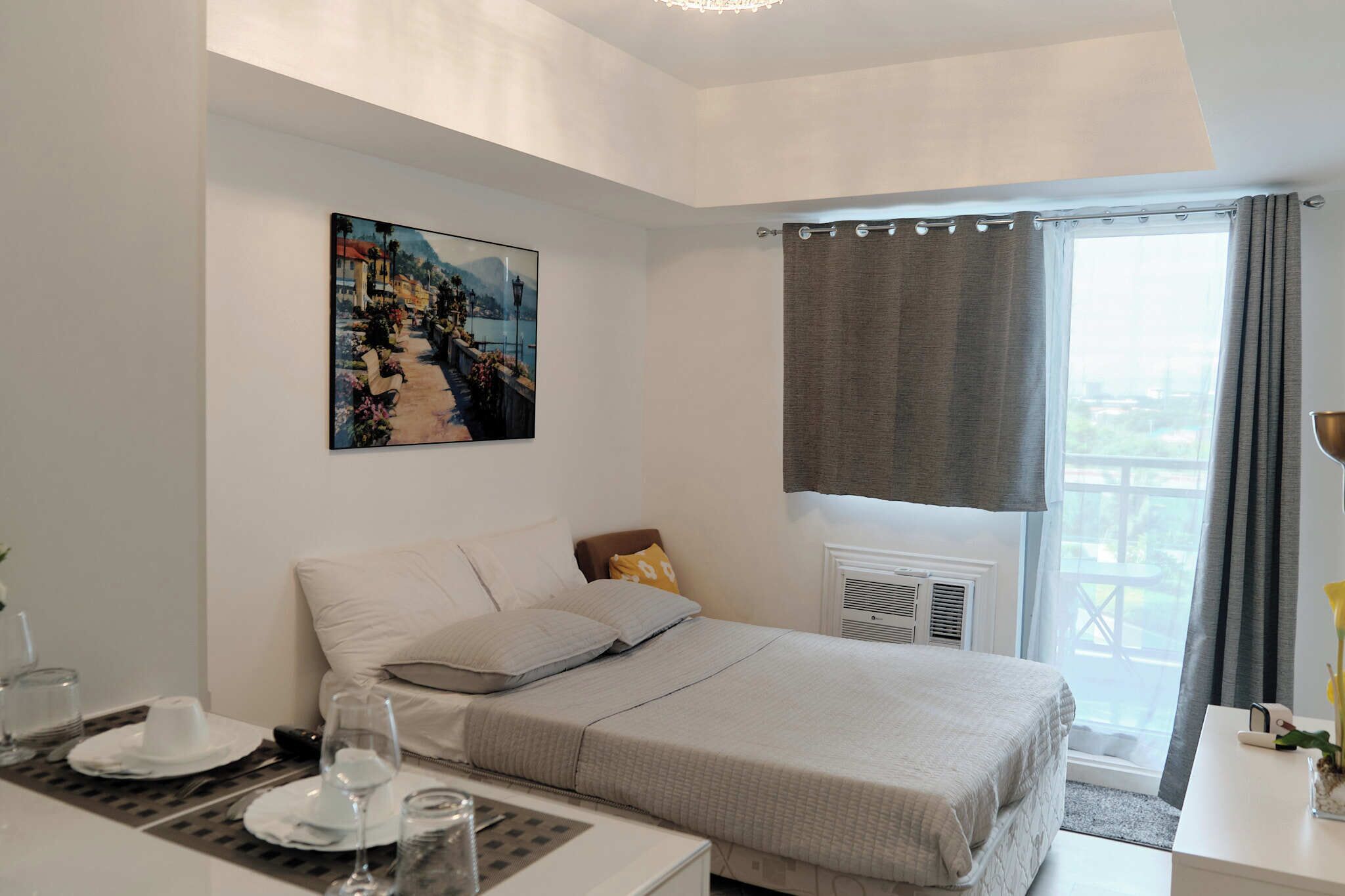 A bedroom with a bed, a table with plates and glasses, a painting on the wall, a window with curtains, and a radiator.