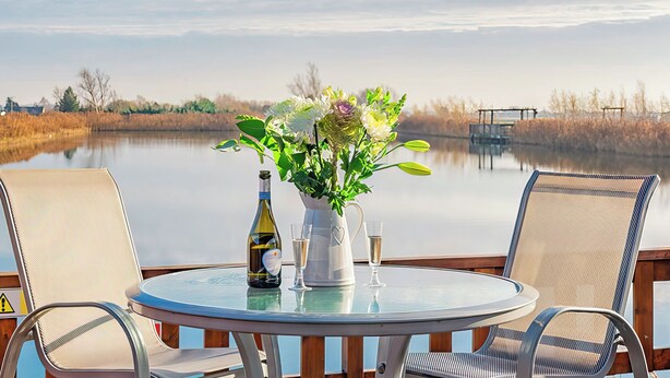 Una mesa redonda con sillas, una botella de vino y un florero con flores en un balcón con vistas a un lago tranquilo.