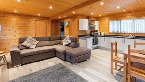 Un acogedor interior de cabaña con un sofá seccional, una mesa de comedor con sillas, una zona de cocina y un techo de madera.