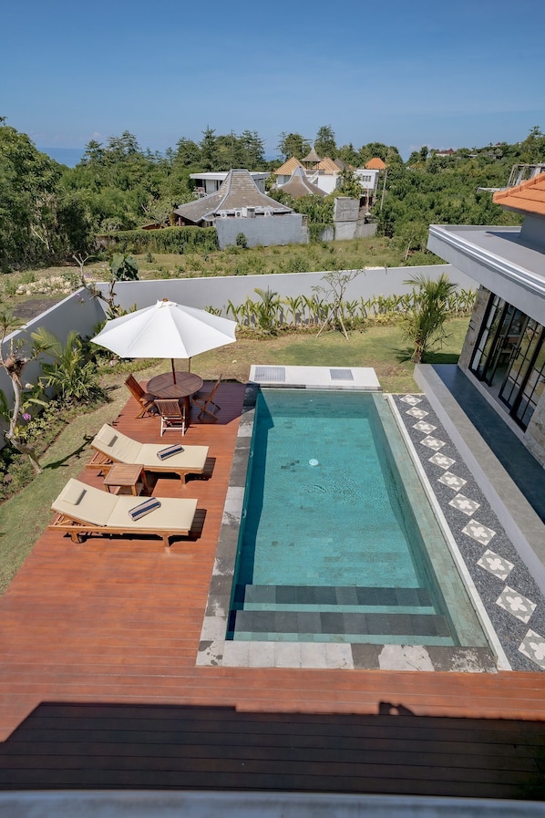 A rectangular swimming pool with a wooden deck, lounge chairs, and a table with an umbrella.