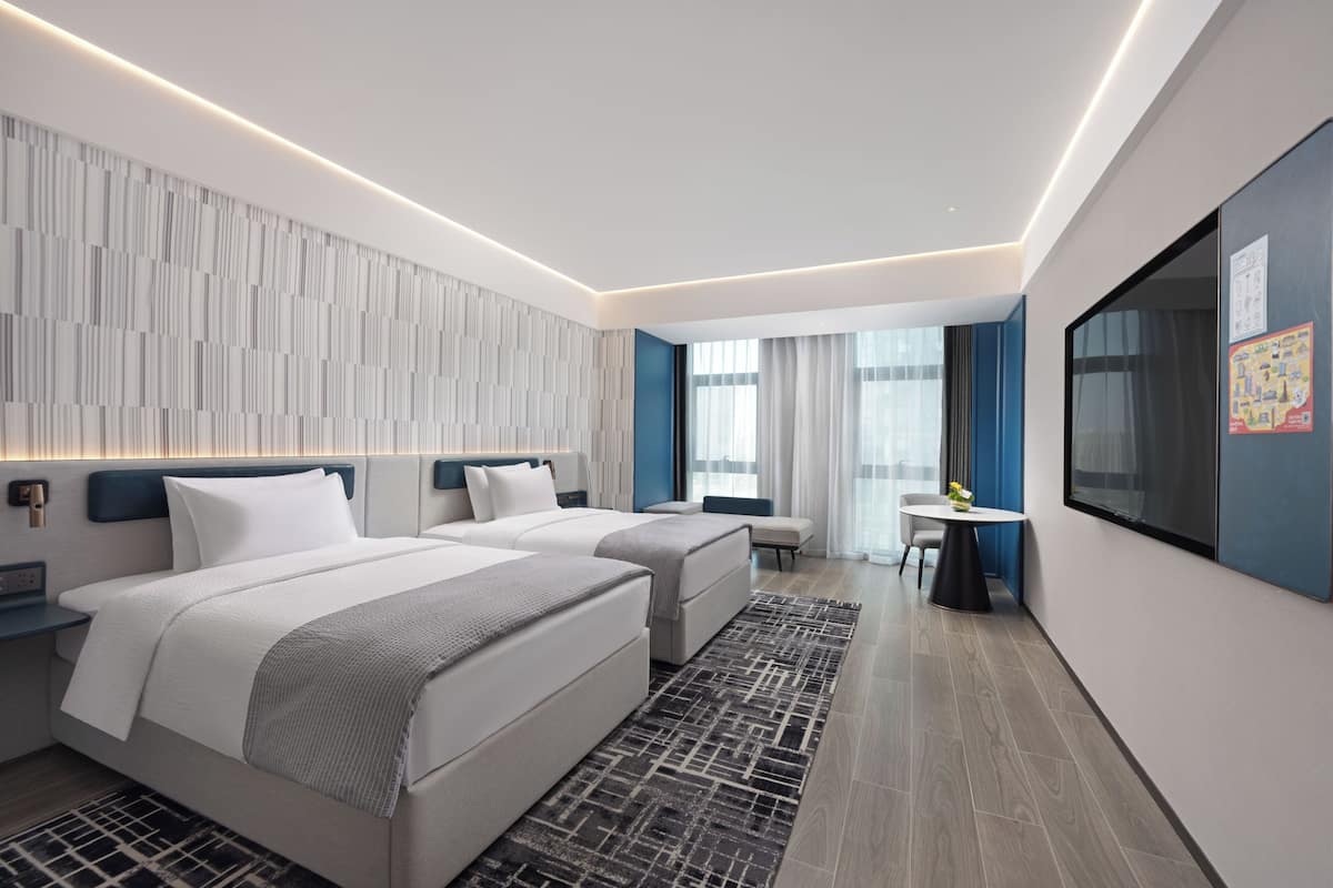 A modern hotel room with two beds, a desk, and a large window.