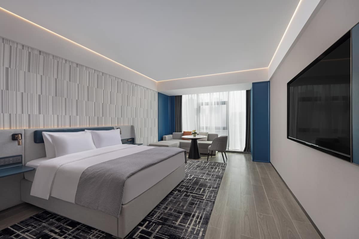 A modern hotel room with a large bed, a desk, a chair, a small table, and a large window.