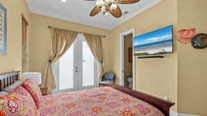 A bedroom with a bed, a ceiling fan, a door leading to a bathroom, and a beach view on the wall. - Vitamin Sea | Easy Beach Living • Community Pool • 3 Min Walk to Beach (South Padre Island)