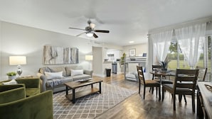 A spacious living area with a sofa, coffee table, and dining table. A ceiling fan is visible, and there is a kitchen area in the background. - Spacious and Stylish Apartment | AT&T Stadium (Arlington)