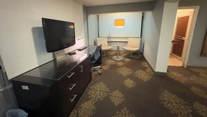Habitació d'hotel amb televisió de pantalla plana, escriptori, dues cadires, taula petita amb làmpada, moqueta amb dibuixos i una porta que dona a una altra habitació.