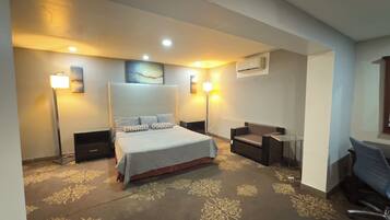 Habitación de hotel con una cama grande, un escritorio, una silla y un sofá. Hay dos mesitas de noche, una lámpara de pie y un equipo de aire acondicionado en la pared.