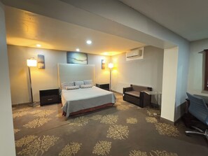 A hotel room with a large bed, a desk, a chair, and a sofa. There are two bedside tables, a floor lamp, and an air conditioning unit on the wall.