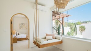 A room with a hanging swing bed, a wooden archway, and a large window with a view of cacti and a building. - New 3BR Chic Pool Villa Near Bingin Beach Uluwatu (Bali)
