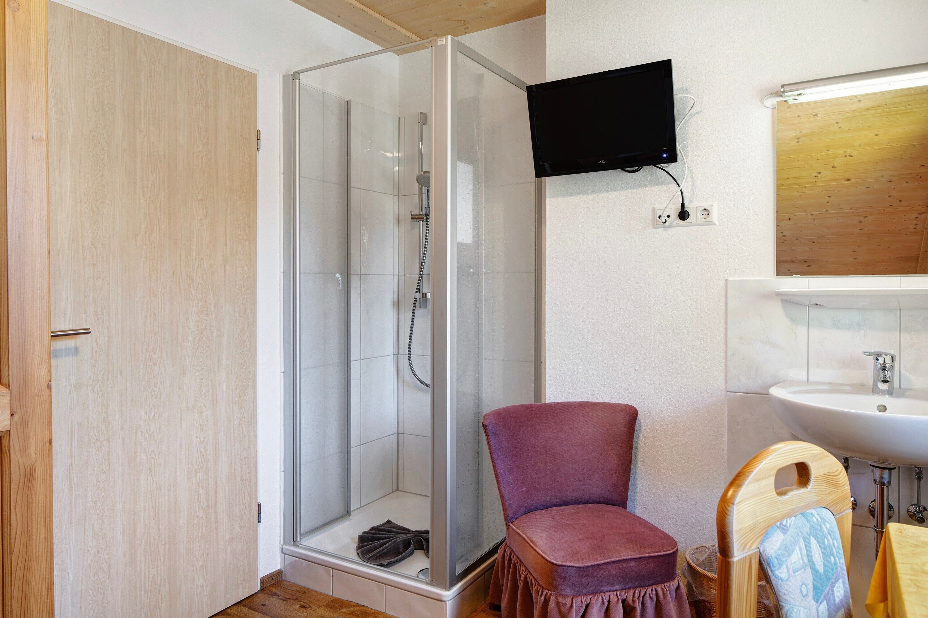 A room with a glass-enclosed shower, a wooden door, a purple chair, a television mounted on the wall, and a sink with a mirror.