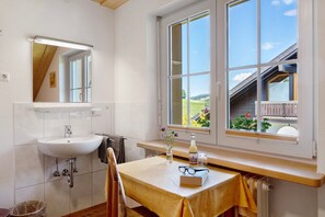 Une salle de bain avec un lavabo, un miroir, une fenêtre et une table sur laquelle se trouvent un livre et des lunettes.