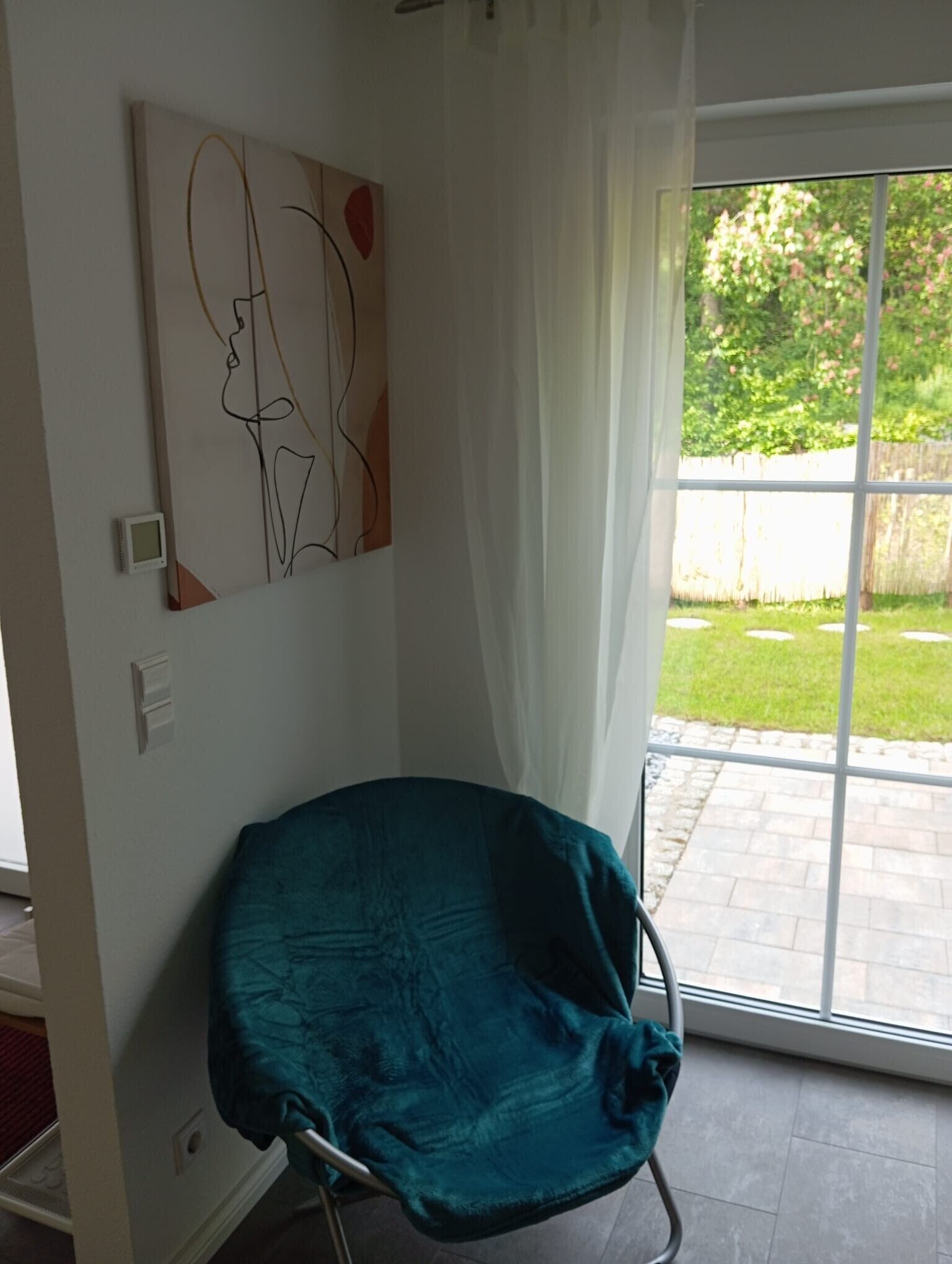 Ein Zimmer mit einem türkisfarbenen Sessel, einem Bild an der Wand und Blick auf einen Garten durch eine Glastür.