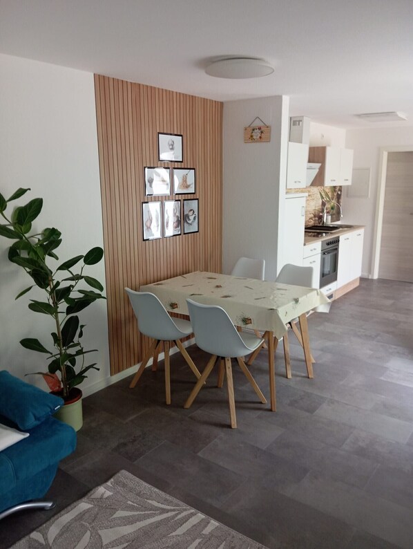 A modern living room with a dining table, chairs, a sofa, a potted plant, and a kitchen area with cabinets and a sink.