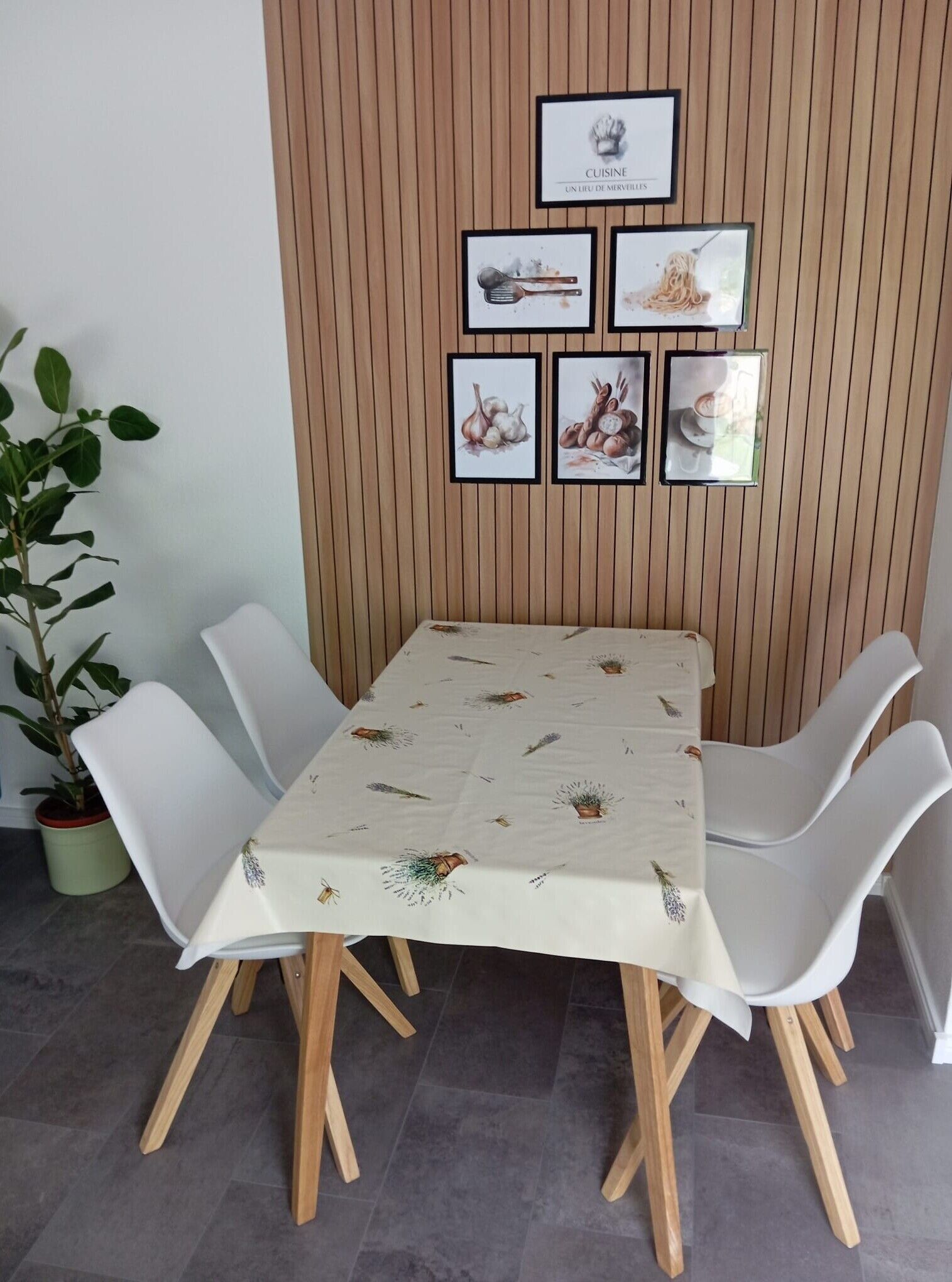 A dining area with a table covered by a tablecloth with plant patterns, surrounded by white chairs, against a wooden wall with framed pictures.