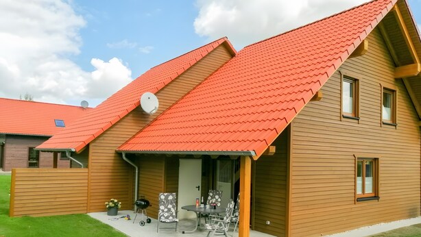 Une maison en bois à deux étages, avec un toit rouge, une terrasse couverte aménagée avec du mobilier d’extérieur et une antenne parabolique.