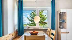 Un comedor con una mesa de madera, sillas, un cuenco de fruta, una lámpara colgante y vistas a árboles a través de una ventana.
