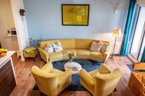 Uma sala de estar moderna com um sofá amarelo, duas poltronas, uma mesa de centro de vidro e uma decoração de cabeça de veado na parede.