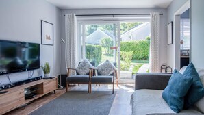 Un salón moderno con un televisor de pantalla plana, un mueble bajo de madera, dos sillones grises, un sofá gris y un amplio ventanal corredera que da a un jardín.